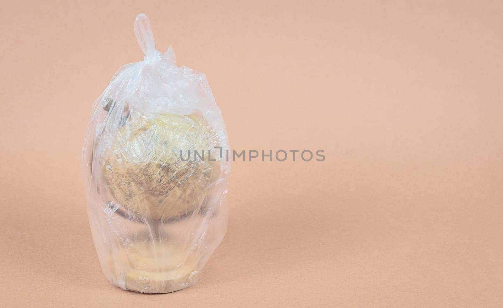 One planet earth globe inside a plastic bag stands on the left on a beige background with copy space on the right, side view close-up.
