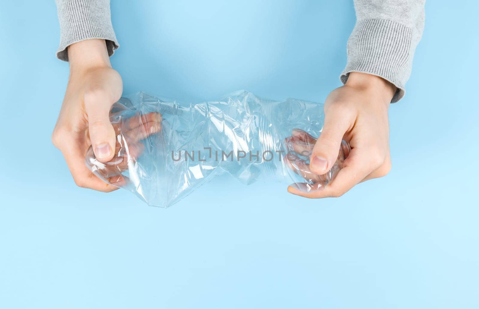 Hands of a young Caucasian man crumpling a transparent plastic bottle on a pastel blue background, close-up, flat lay closeup. The concept is environmentally friendly, sorting and recycling waste, protecting nature.