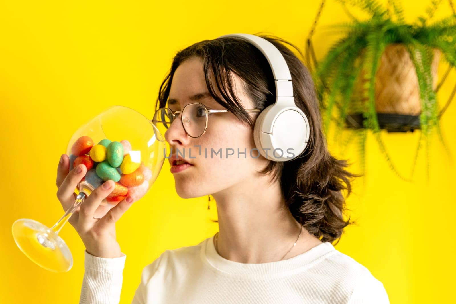 One young Caucasian beautiful teenage girl in glasses and headphones holds a wine glass with Easter marble decorative eggs standing and looking to the side on a yellow background with a hanging palm flower on a spring day in the room, side view close-up.