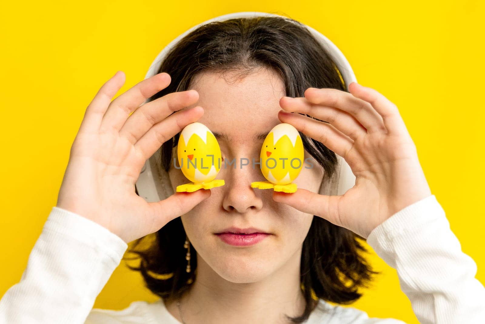 Portrait of one young Caucasian beautiful happy teenage girl holding with her fingers two Easter decorative chickens at eye level, standing on a yellow background on a spring day in the room, side view close-up.