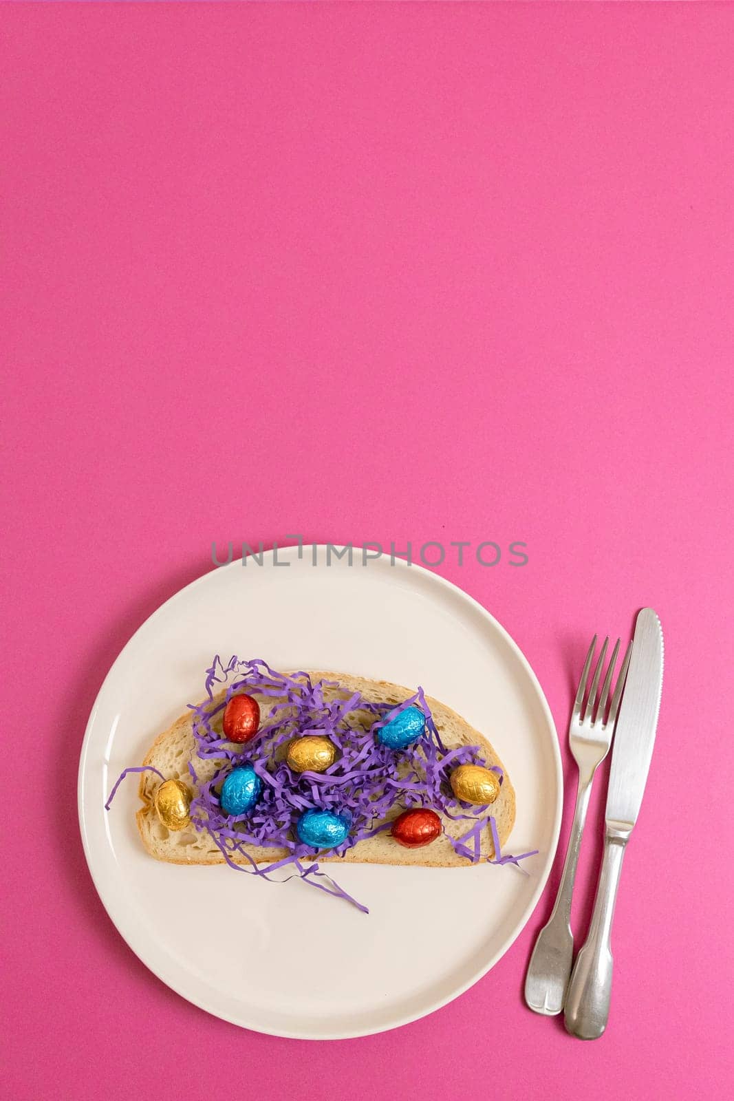 One sandwich with decorative lilac paper hay with chocolate Easter eggs in shiny multi-colored wrappers on a tray with a fork and knife lie from below on a pink background with copy space on top, flat lay close-up. Creative Easter food concept.
