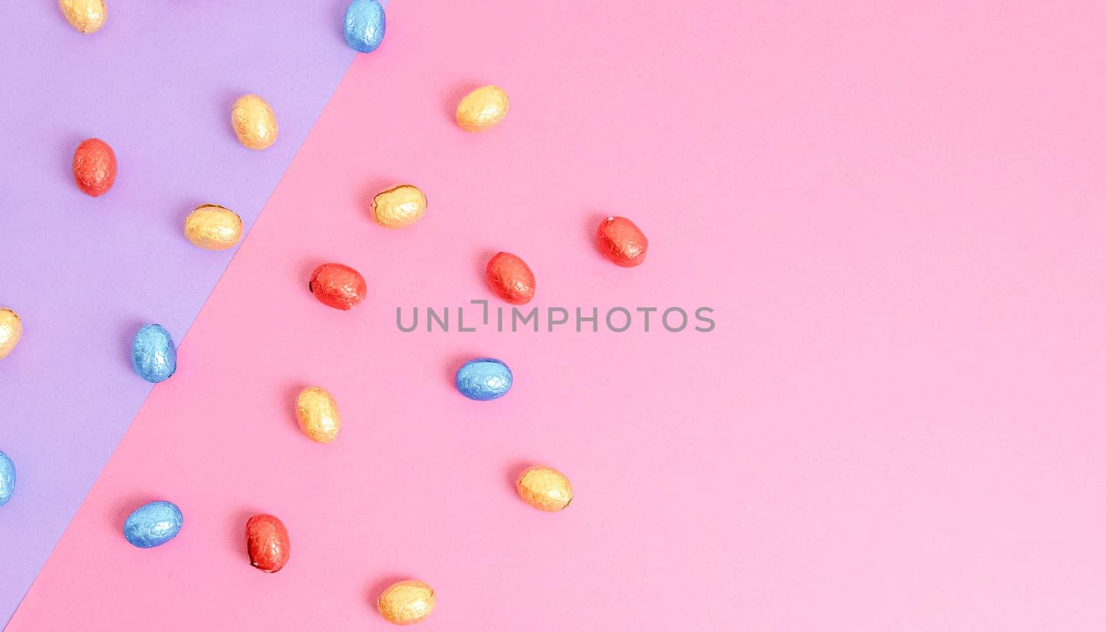 Scattered chocolate Easter eggs in shiny multi-colored wrappers lie on the left on a lilac-pink background with copy space on the right, flat lay close-up.Easter food concept.