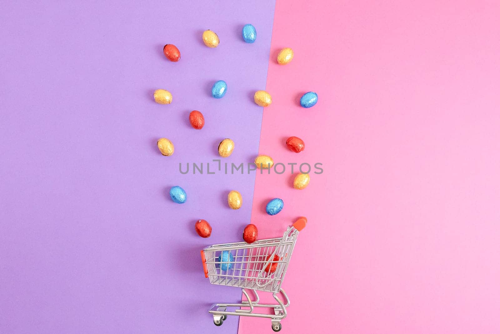 Chocolate Easter eggs in shiny multi-colored wrappers spill out of a mini shopping cart, lie in the center on a lilac-pink background with copy space on the sides, flat lay close-up. Easter shopping concept.