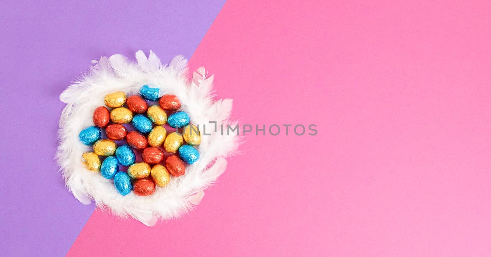 Chocolate Easter eggs in shiny multi-colored wrappers inside a wreath of white berets lie on the left on a lilac-pink background with copy space on the right, flat lay close-up. Easter shopping concept.
