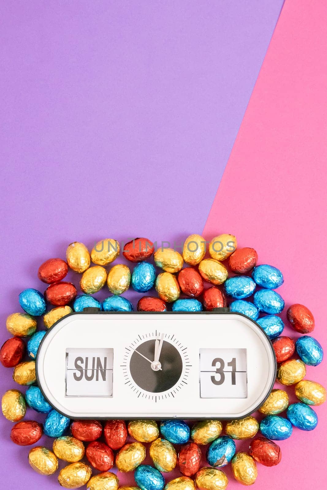 One alarm clock with the date and time of Easter 2024: Sunday March 31st with chocolate Easter eggs in colorful shiny wrappers lie from below on a lilac pink background with copy space on top, flat lay close-up. Easter candy concept, holiday time.