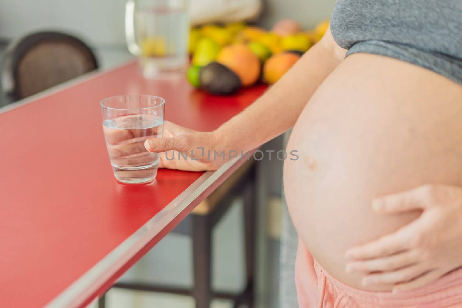 Embracing the vital benefits of water during pregnancy, a pregnant woman stands in the kitchen with a glass, highlighting hydration's crucial role in maternal well-being.