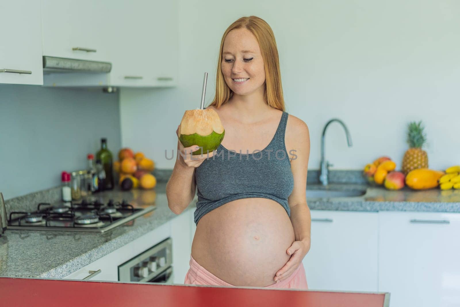 Quenching her pregnancy thirst with a refreshing choice, a pregnant woman joyfully drinks coconut water from a coconut in the kitchen, embracing natural hydration during this special journey.