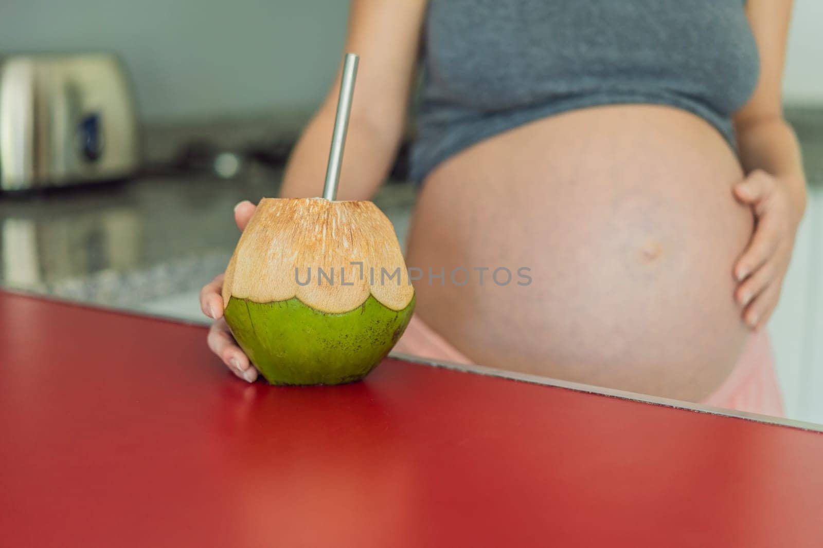 Quenching her pregnancy thirst with a refreshing choice, a pregnant woman joyfully drinks coconut water from a coconut in the kitchen, embracing natural hydration during this special journey by galitskaya