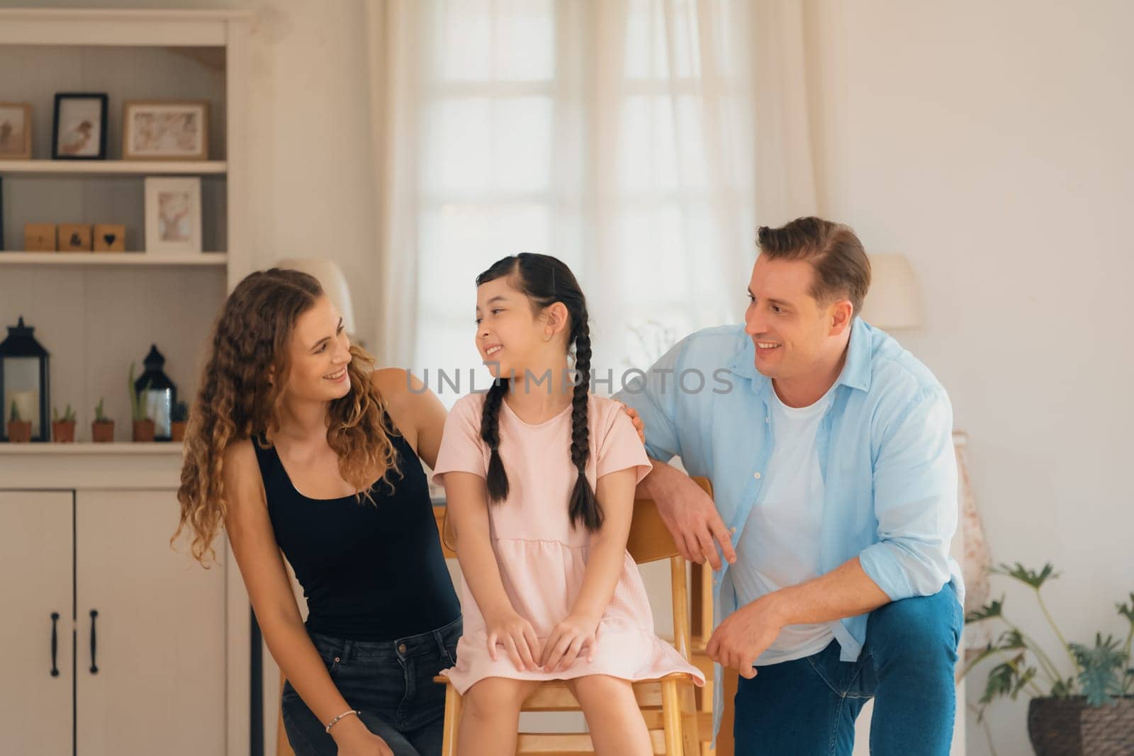 Happy family portrait with lovely little girl smiling and looking at camera, lovely and cheerful parent and their daughter sitting together in living room at home with warm daylight. Synchronos