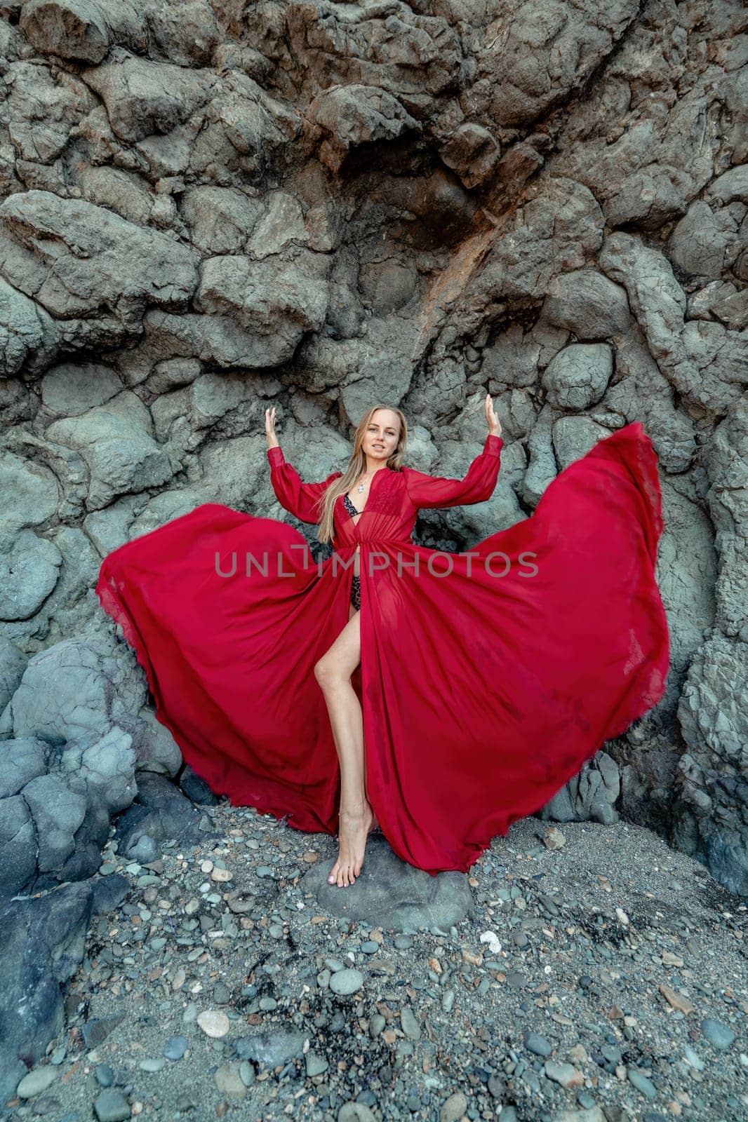 Red dress rocks woman. A blonde with flowing hair in a long flowing red dress stands near a rock of volcanic origin. Travel concept, photo session at sea by Matiunina