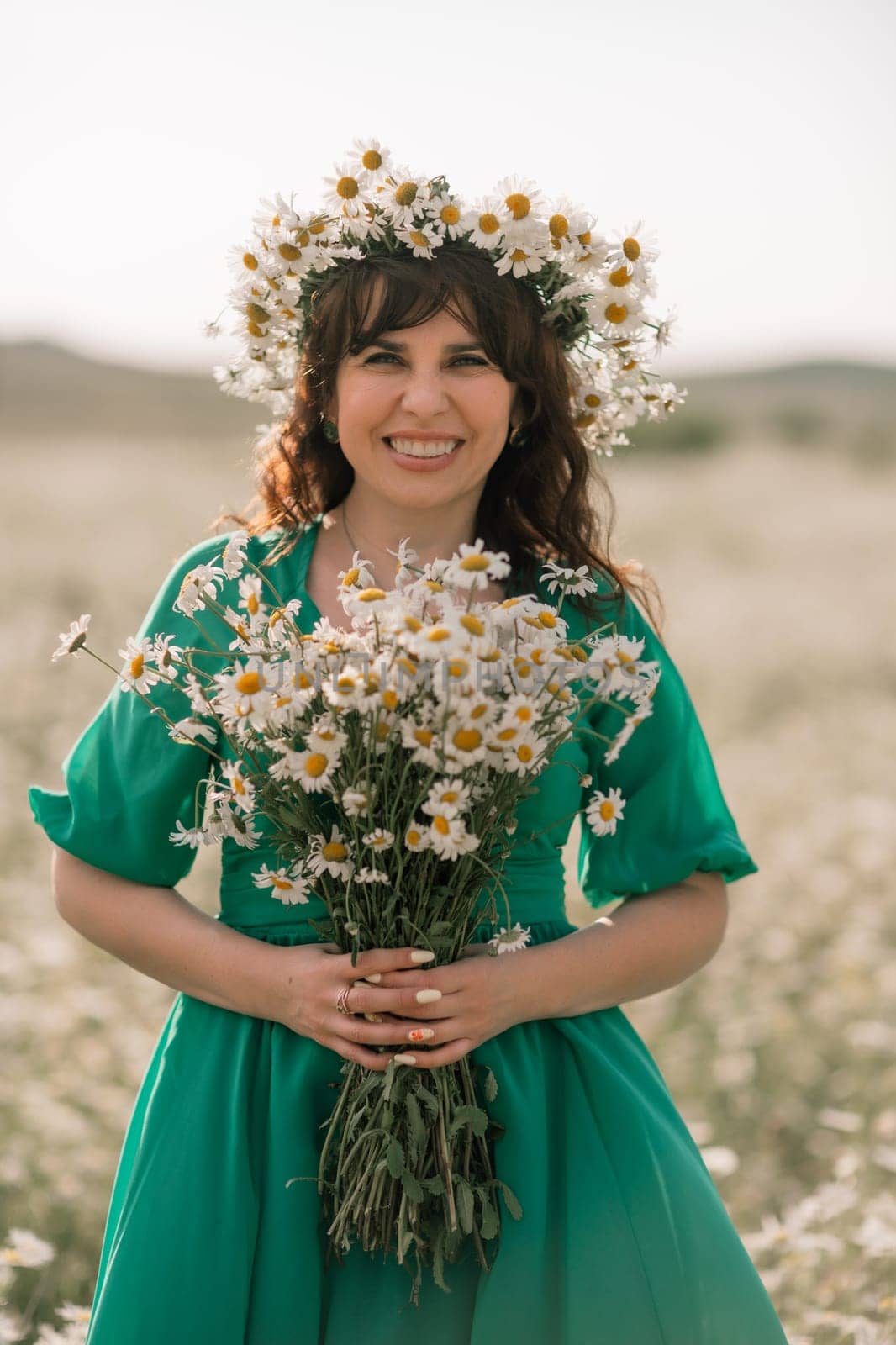 Happy woman in a field of daisies with a wreath of wildflowers on her head. woman in a green dress in a field of white flowers. Charming woman with a bouquet of daisies, tender summer photo.