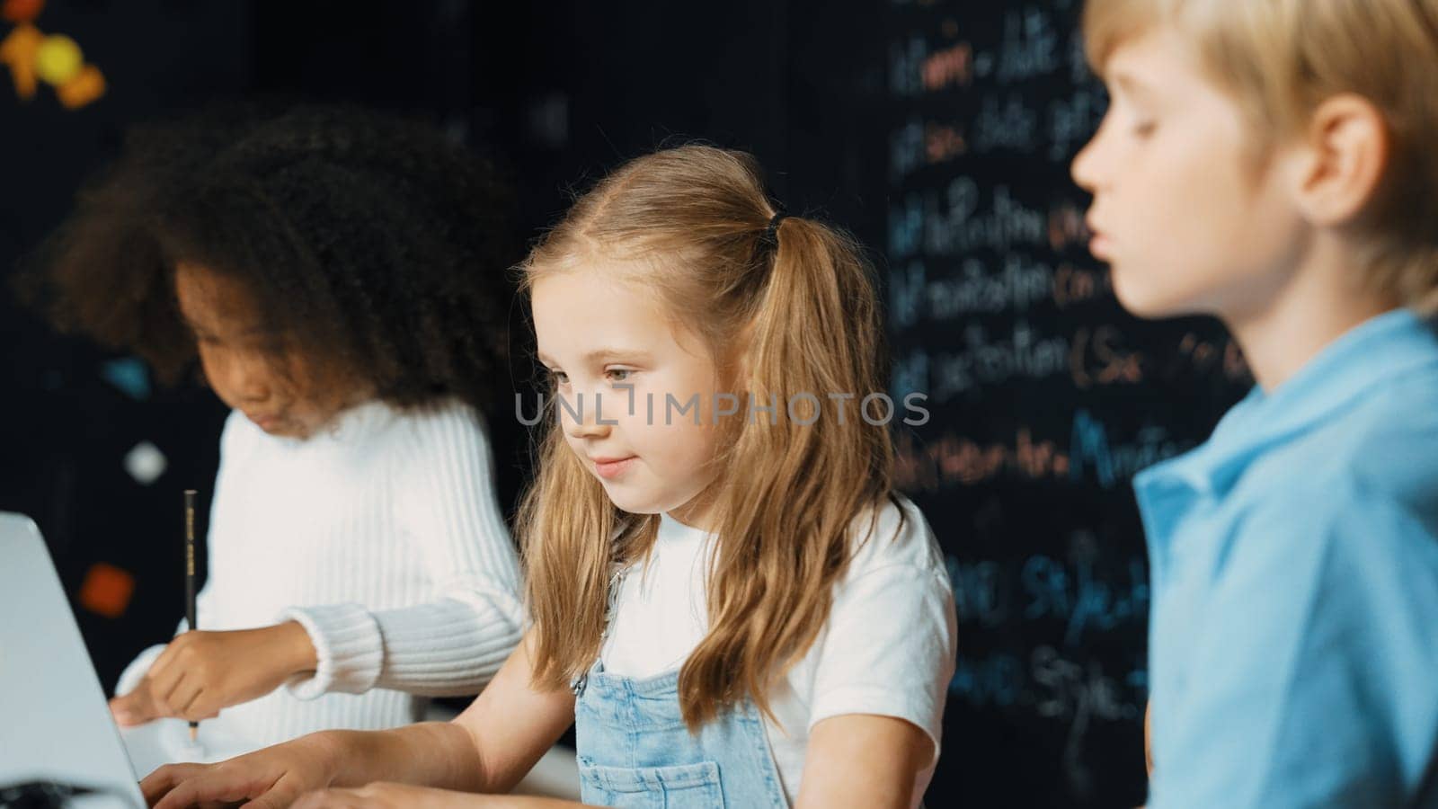 Smart girl using laptop programing engineering code with diverse friends ay table with microchip and main board scatter around in STEM classroom at blackboard written with coding prompt. Erudition.
