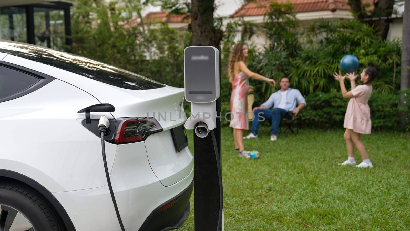 Electric vehicle recharge from home charging station on background of happy and playful family playing together. EV car using alternative and sustainable energy for better future Synchronos