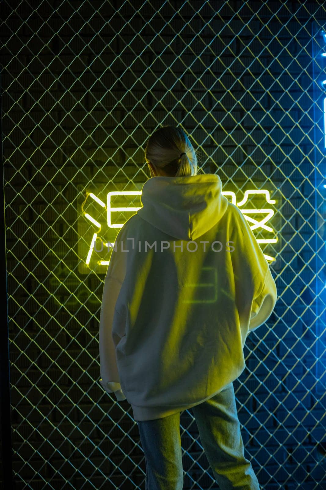 Beautiful blonde woman in white hoodie and blue jeans posing against neon background by Freeman_Studio