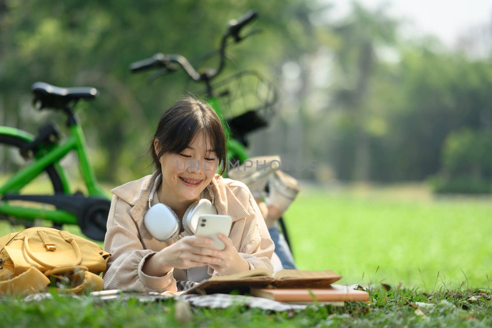 Happy young female using mobile phone while lying on grass near her bicycle in city park.