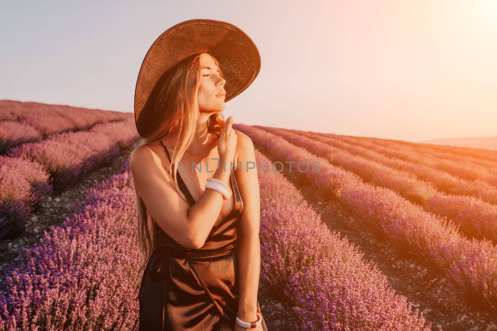 Close up portrait of young beautiful woman in a white dress and a hat is walking in the lavender field and smelling lavender bouquet.