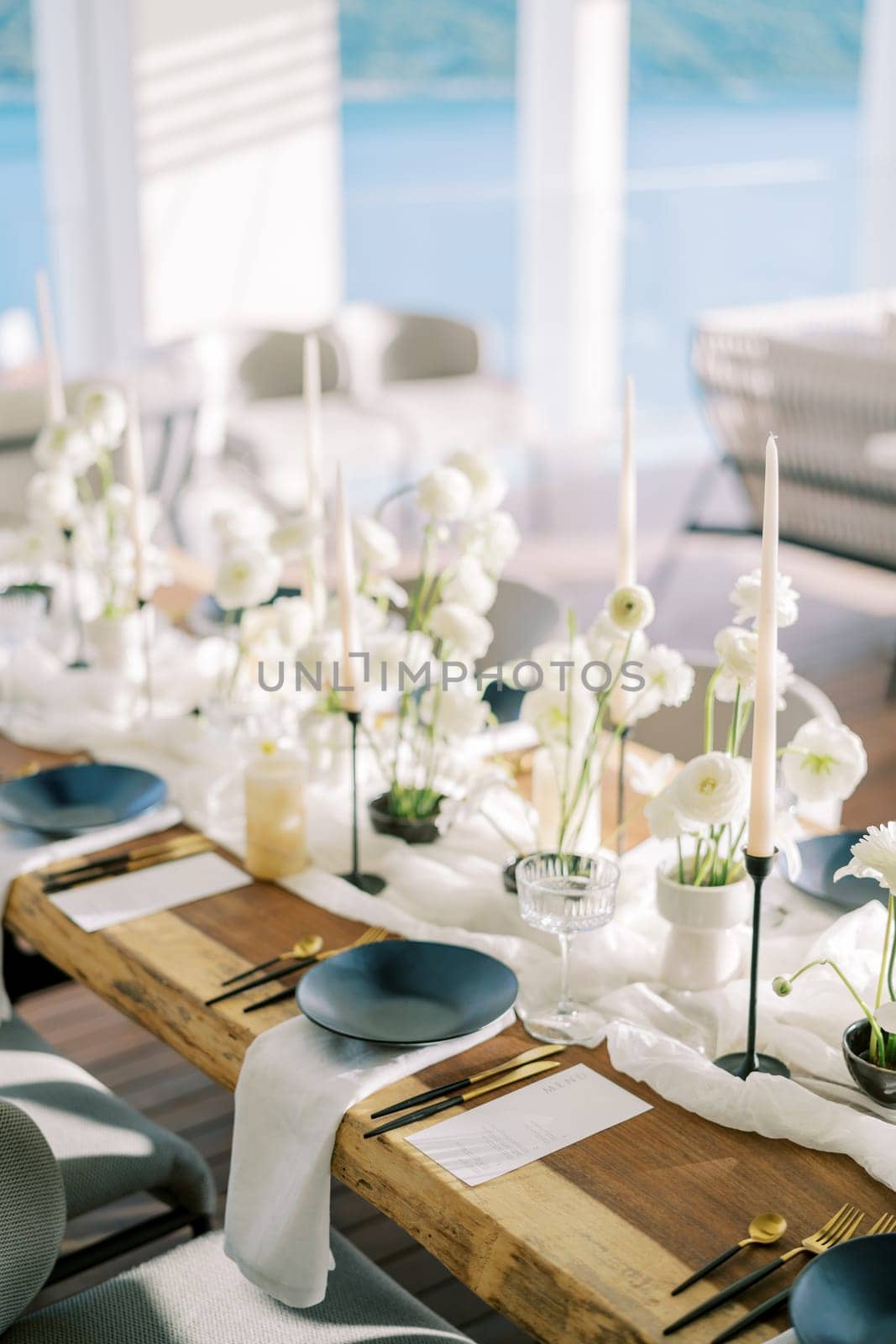 Black plates on white napkins stand next to gold cutlery and menus on a holiday table with white flowers by Nadtochiy