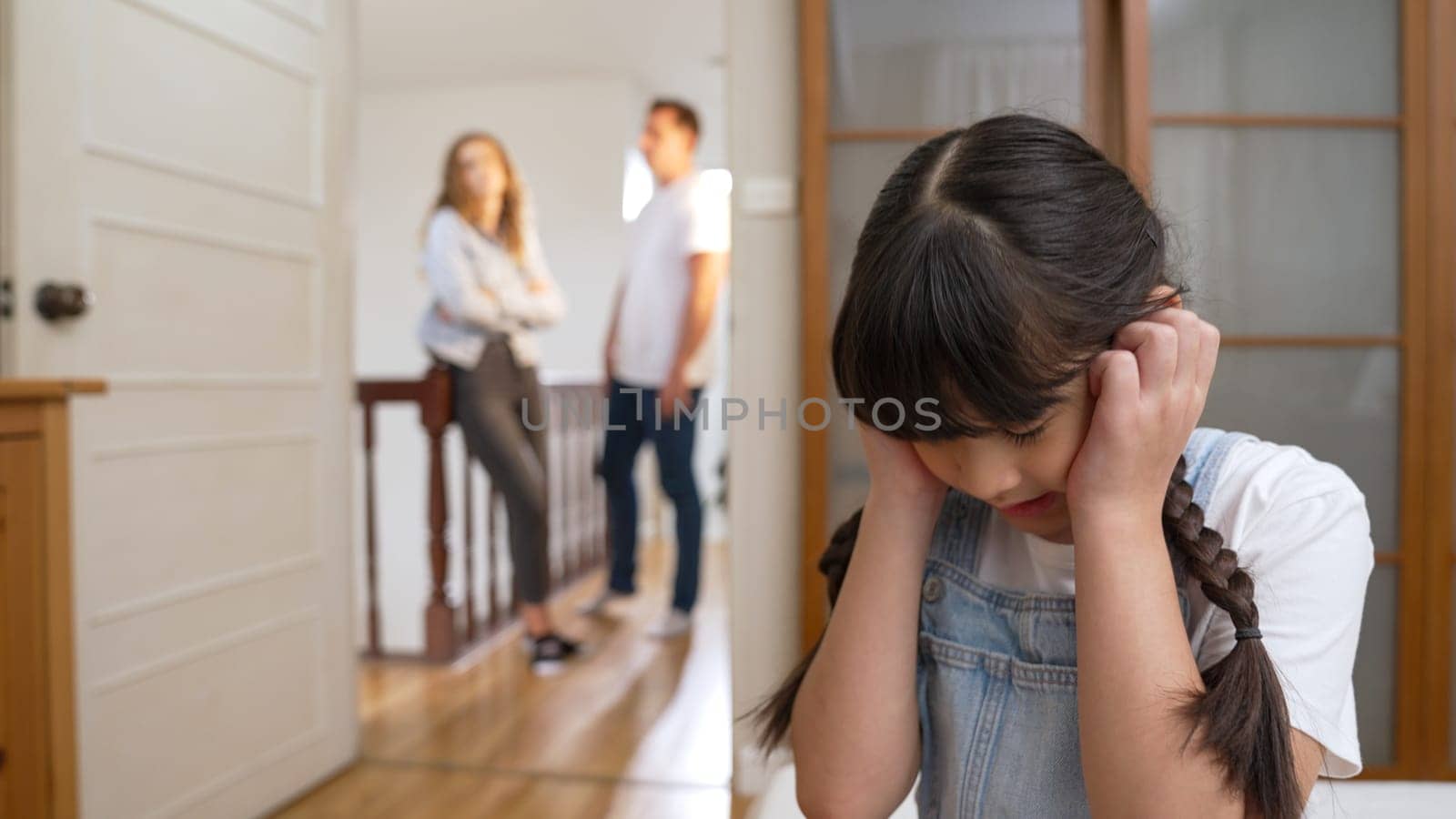 Stressed and unhappy young girl huddle in corner, cover her ears blocking sound of her parent arguing in background. Domestic violence at home and traumatic childhood develop to depression. Synchronos