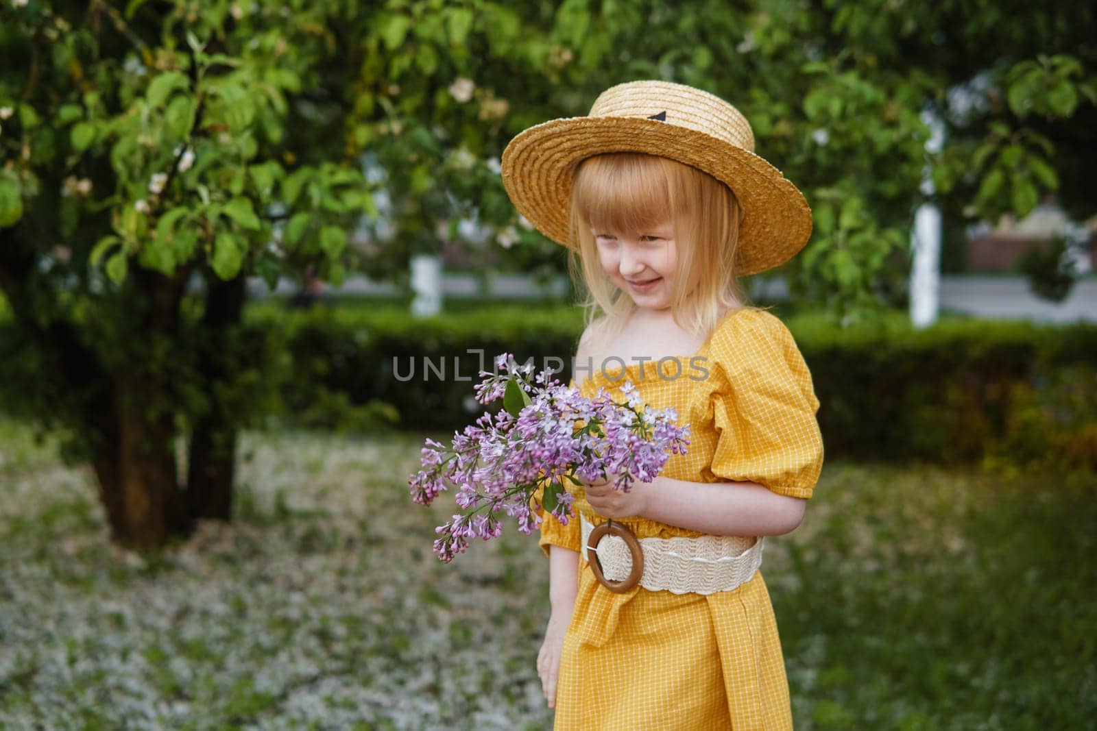 A little girl in a yellow dress and straw hat wearing a bouquet of lilacs. A walk in a spring park, blossoming lilacs