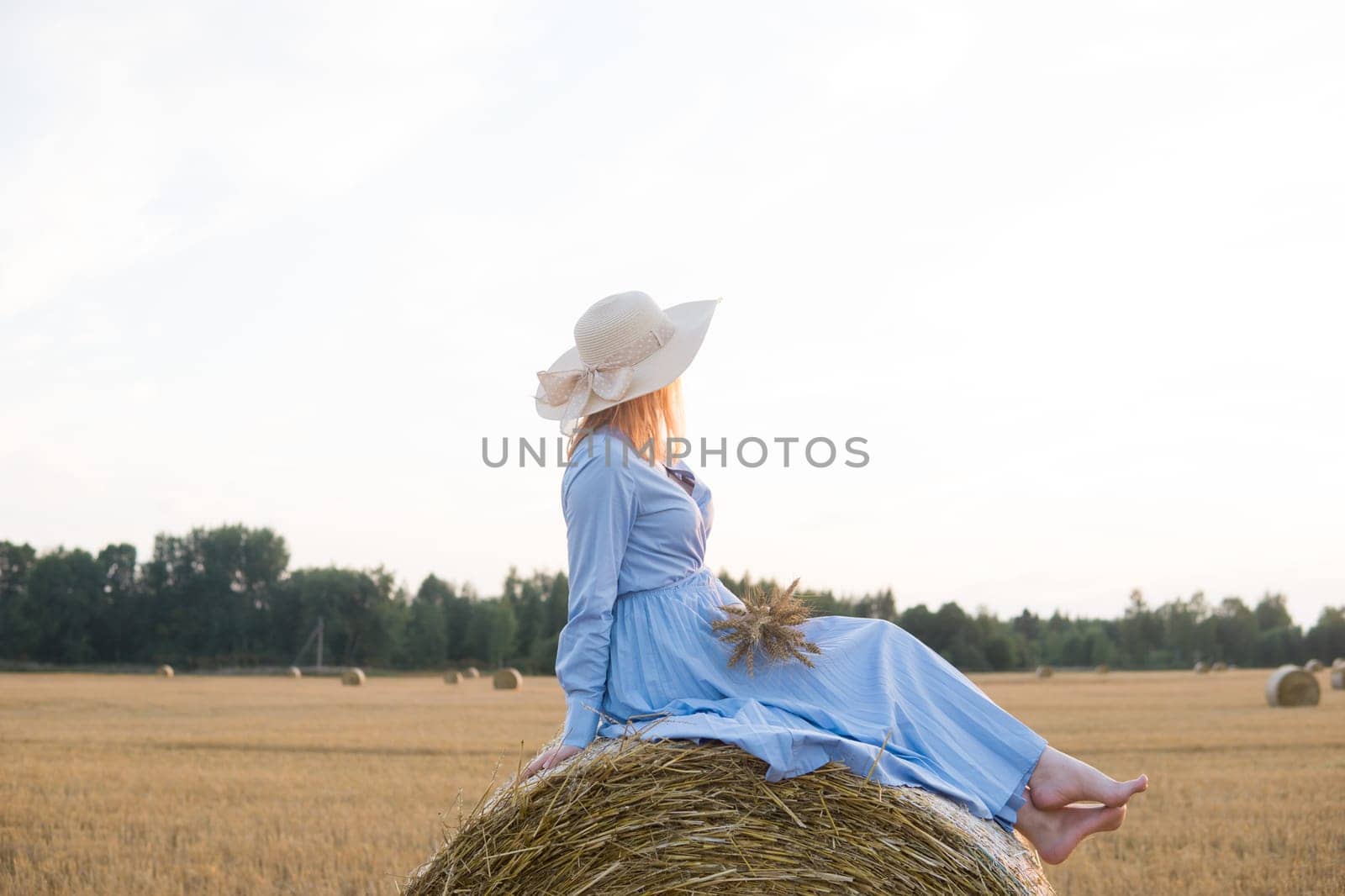 A red-haired woman in a hat and a blue dress walks in a field with haystacks