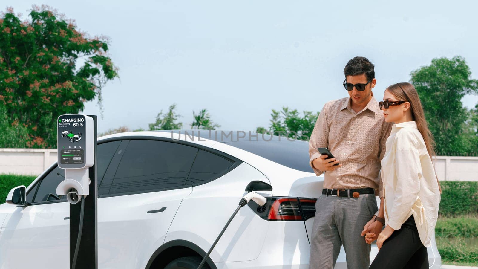 Young couple use smartphone to pay for electricity at public EV car charging station green city park. Modern environmental and sustainable urban lifestyle with EV vehicle. Panorama Expedient