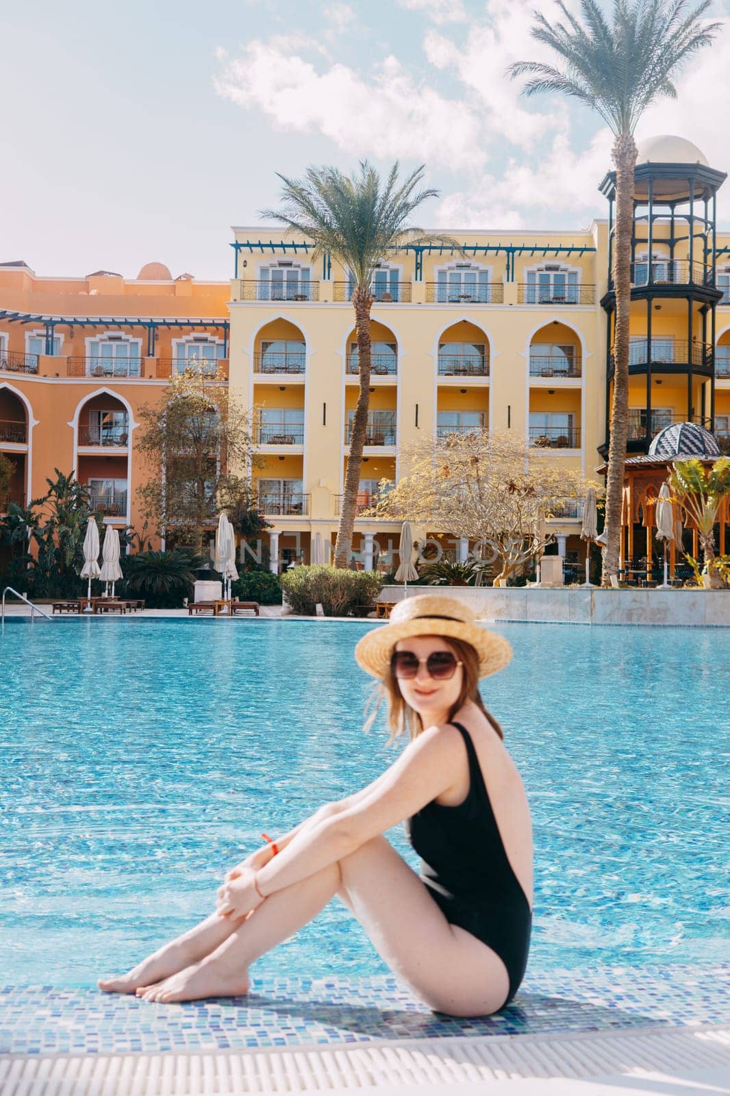 A woman in a black swimsuit, hat and sunglasses is sitting by the pool. The concept of a luxury summer vacation at the hotel.