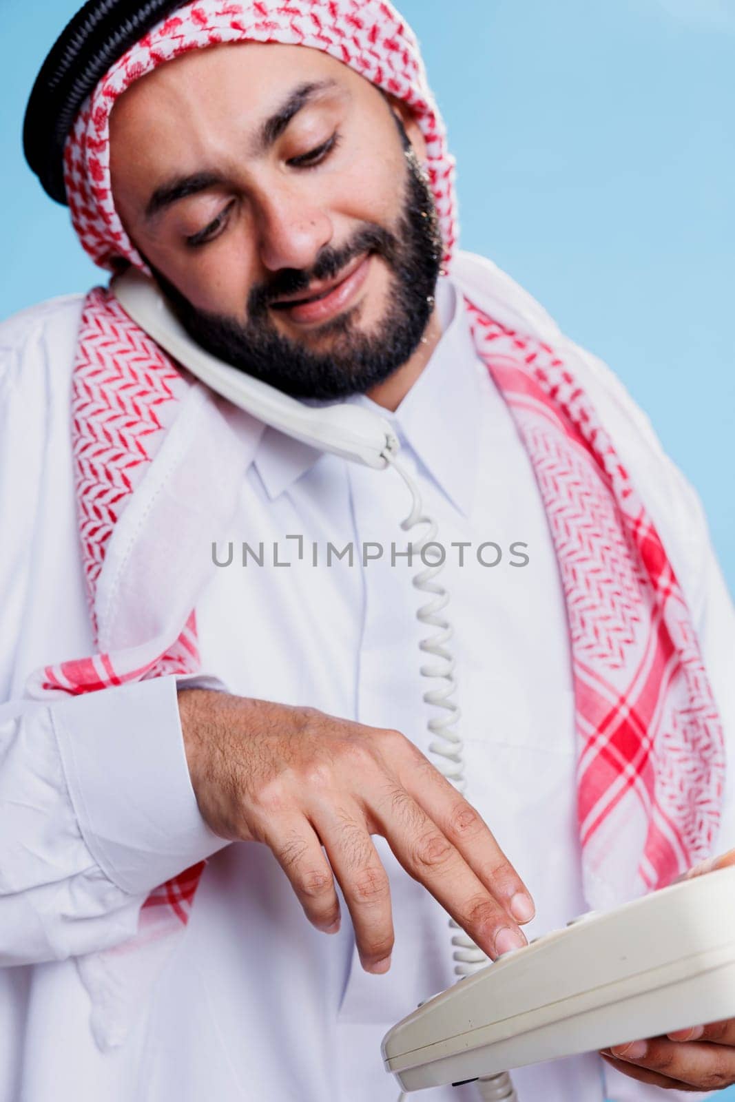 Muslim man wearing traditional headscarf dialing number on landline phone. Arab person dressed in cultural islamic clothes holding retro telephone and making call closeup