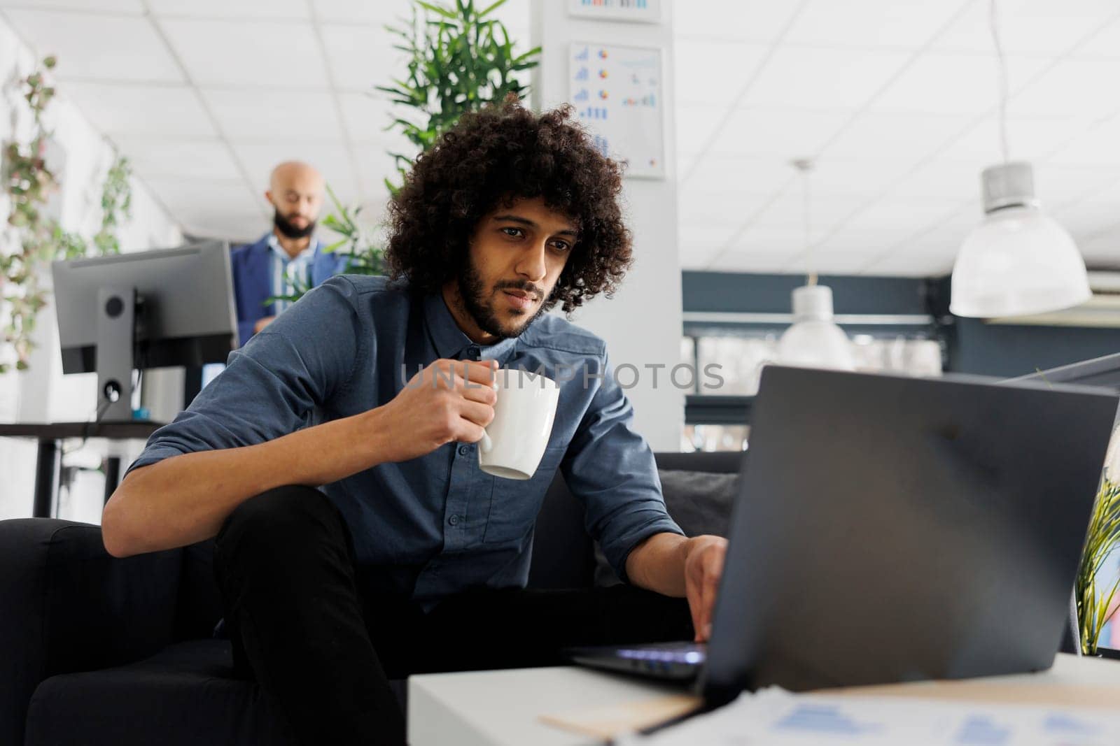 Young arab comapny employee drinking coffee and using corporate laptop in business office. Executive manager holding tea mug and solving work tasks on computer in coworking space