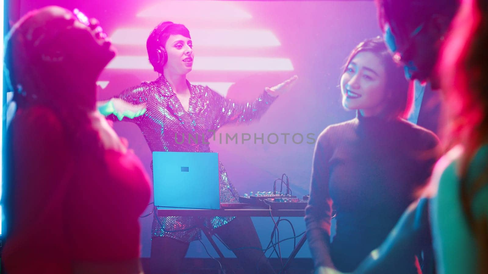 Funky female DJ mixing sounds on stage at club, dancing with group of friends at audio station. Young woman jumping with diverse people, enjoying night out at nightclub party. Tripod shot.