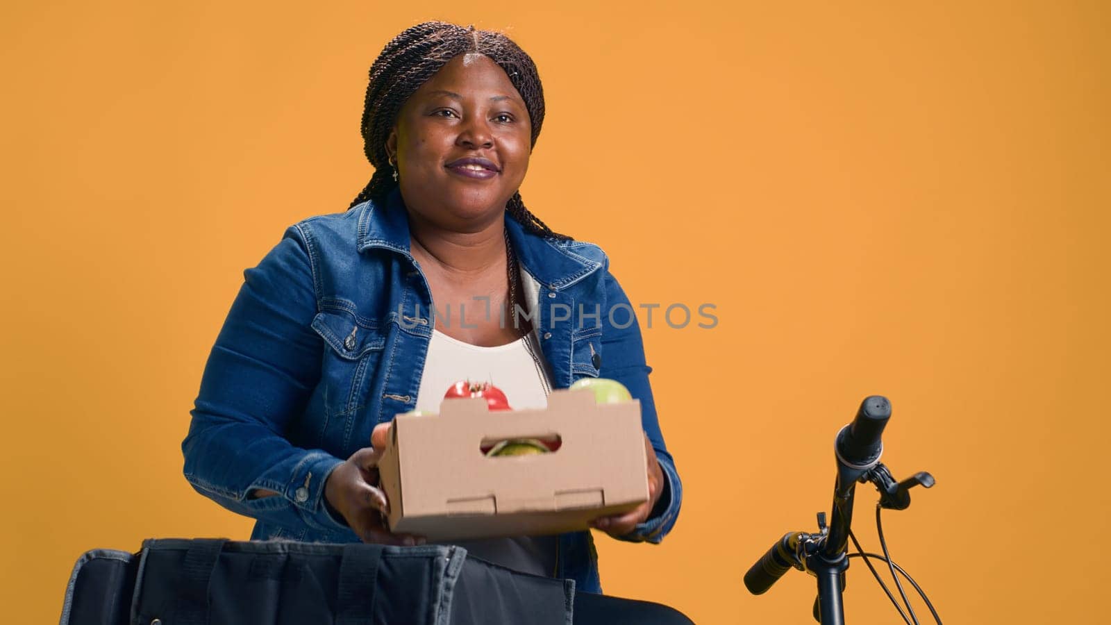 Bicycle is being used by helpful delivery woman to deliver fresh fruits on-demand. Efficient courier service provided by pleasant african american bike messenger with basket of scrumptious vegetables.