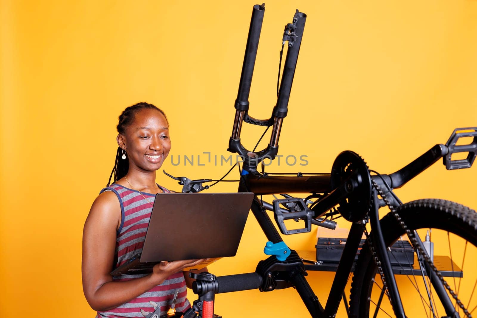 Female cyclist of african american ethnicity fixes damaged bike using toolkit and laptop to research solutions. Using minicomputer and professional equipment, black woman is repairing broken bicycle.