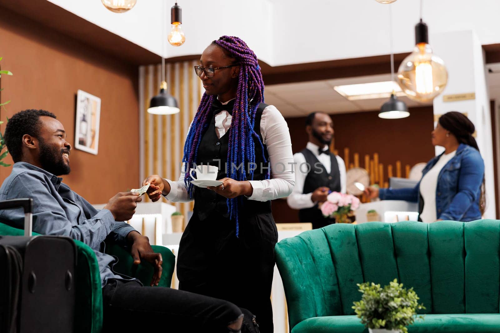Hotel waitress holding cup of freshly brewed coffee taking cash payment from guest while serving hot drink. Happy African American man traveler ordering espresso while waiting in lobby for check-in