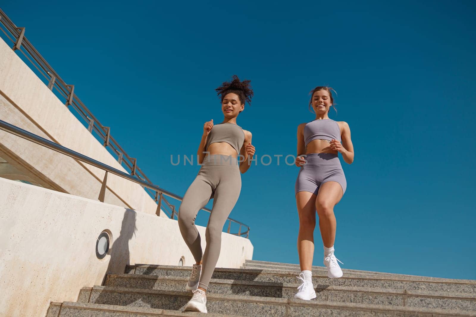 Two active female athlete friends in sportswear running on steps outdoors on a sunny day by Yaroslav_astakhov