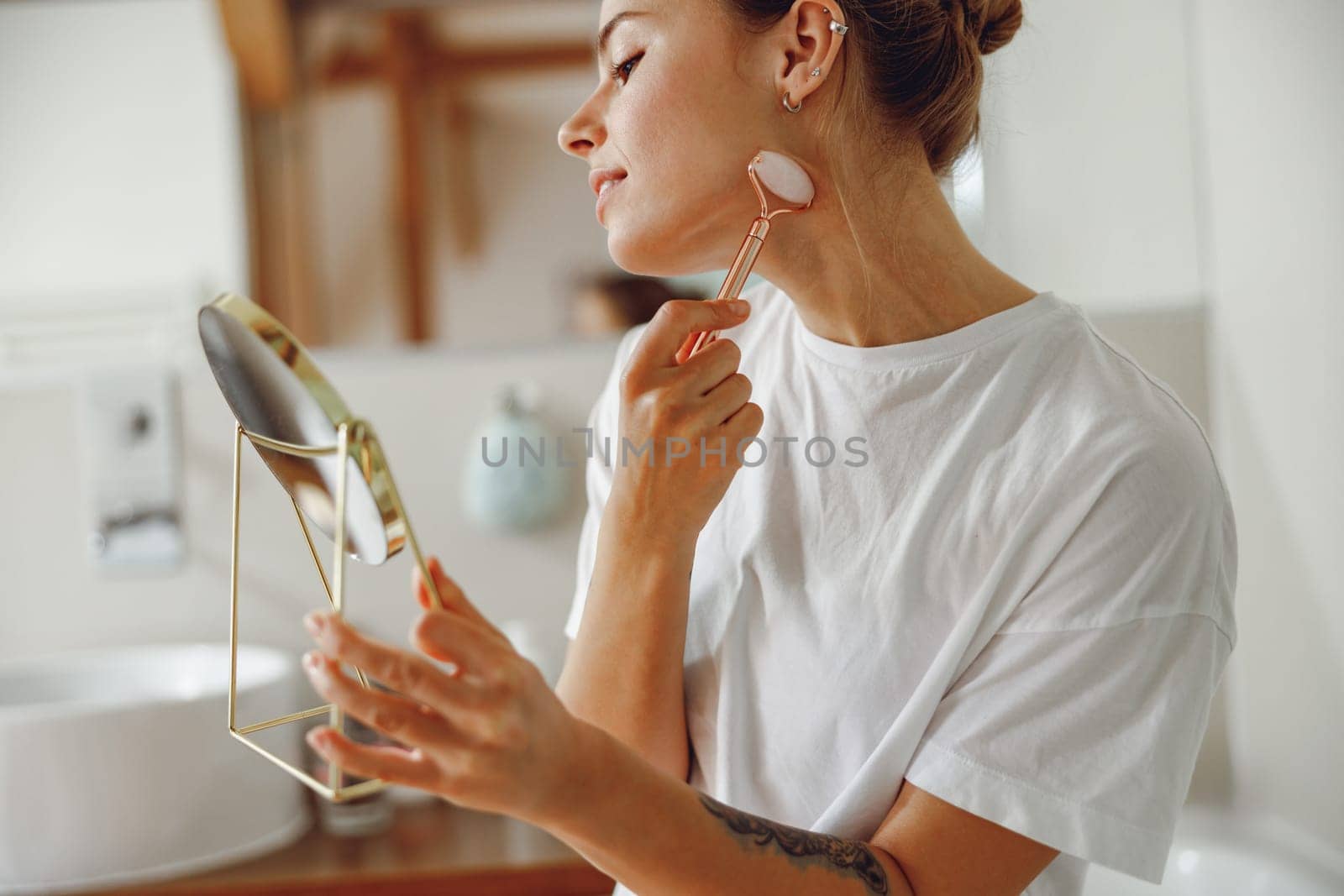 Smiling woman massaging her face with jade roller massager and looking at round cosmetic mirror