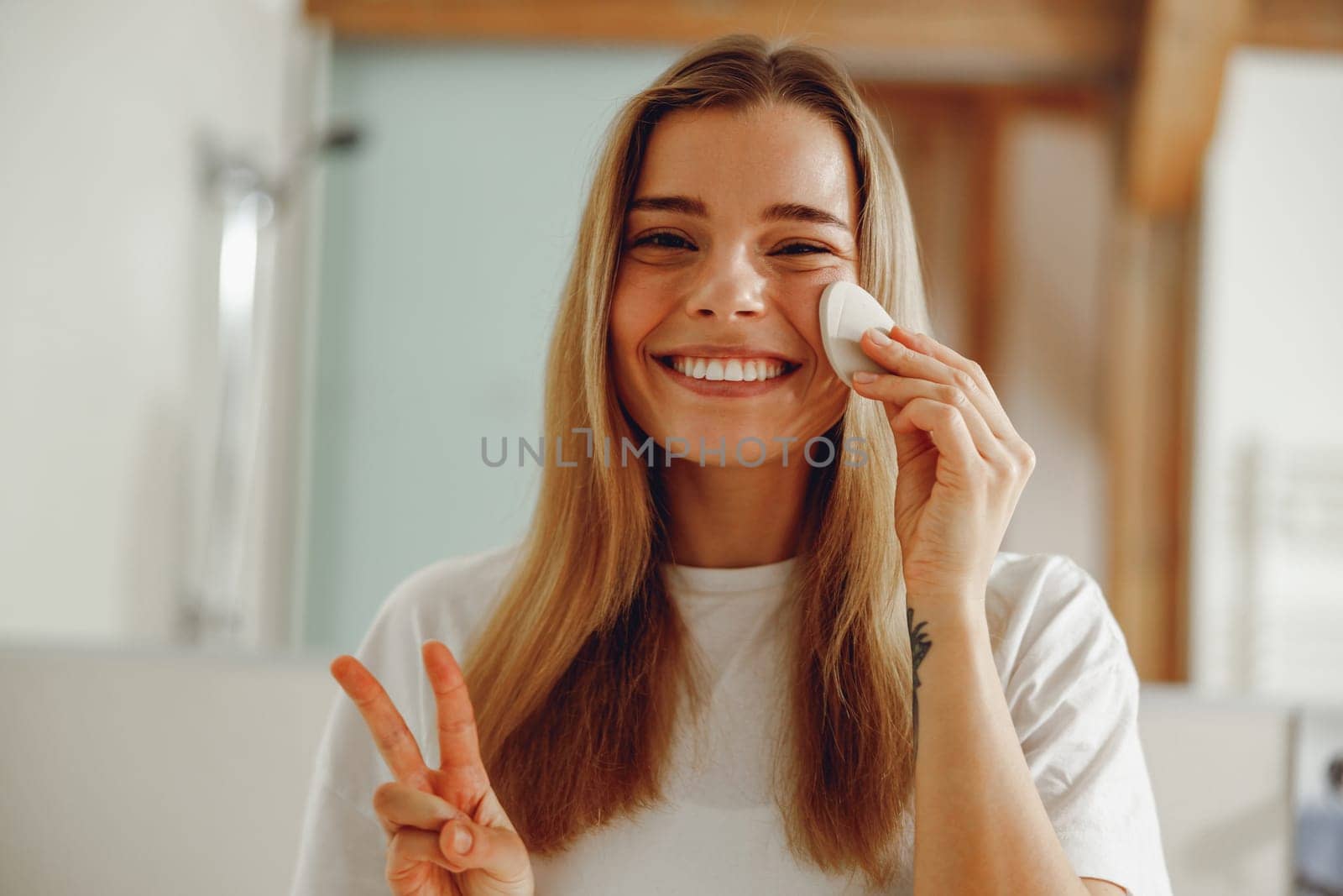 Woman wiping her face with cotton pad in the bathroom and showing sign peace. Skin care concept by Yaroslav_astakhov
