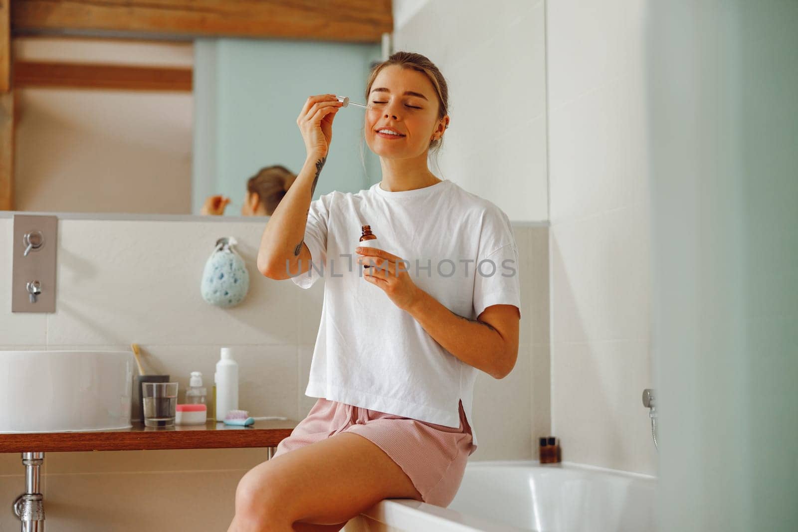 Beautiful young woman applying cosmetic serum onto her face in bathroom. Facial Skincare