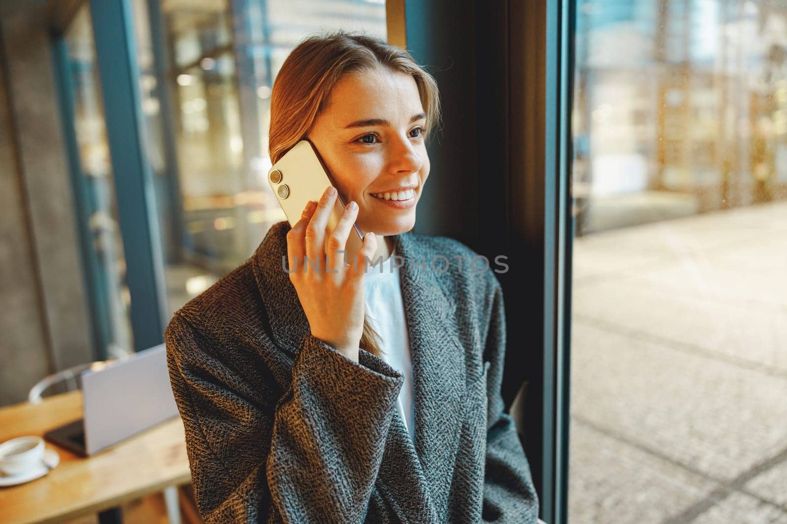 Smiling businesswoman is talking phone with colleagues while standing in cafe near window