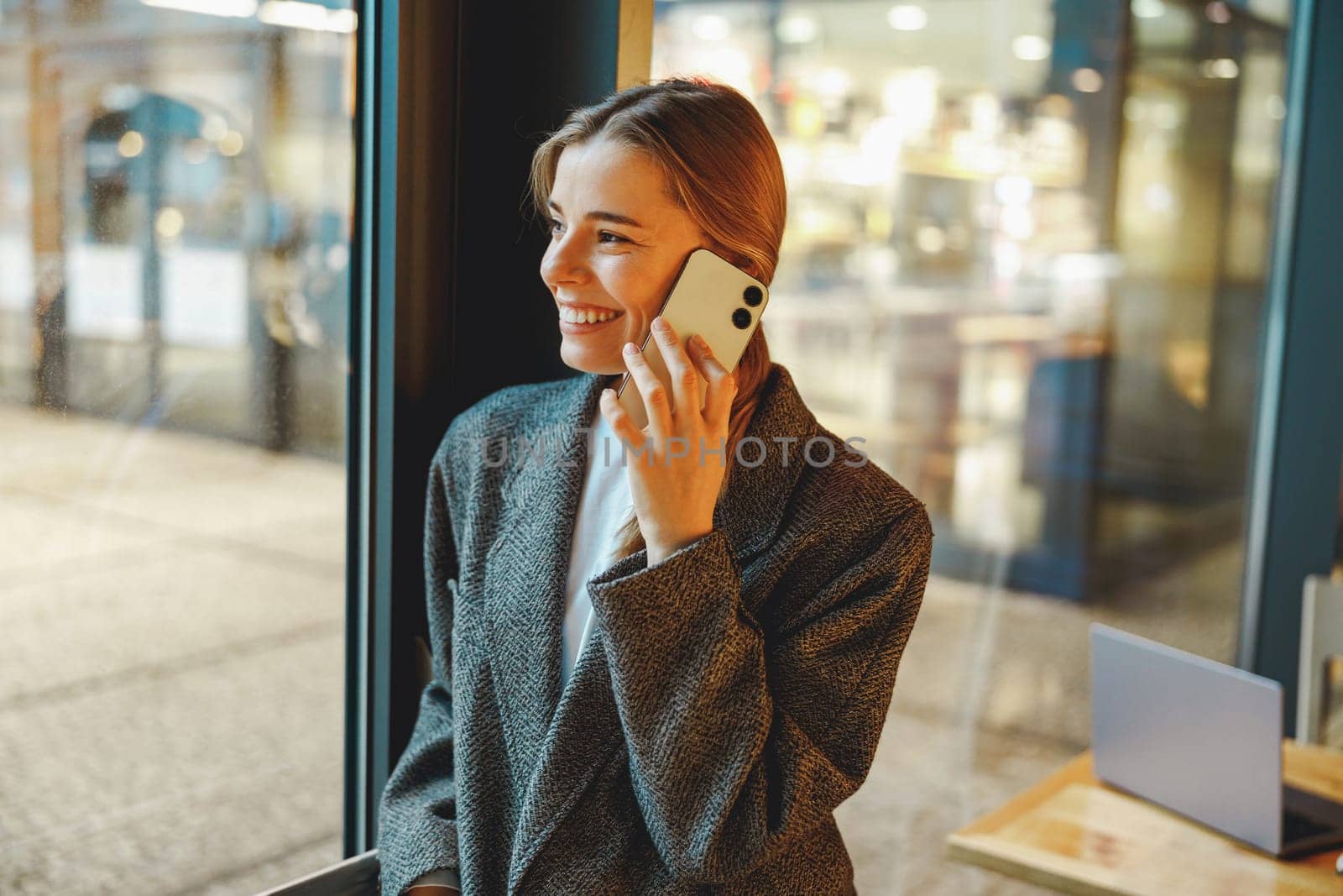 Smiling businesswoman is talking phone with colleagues while standing in cafe near window by Yaroslav_astakhov