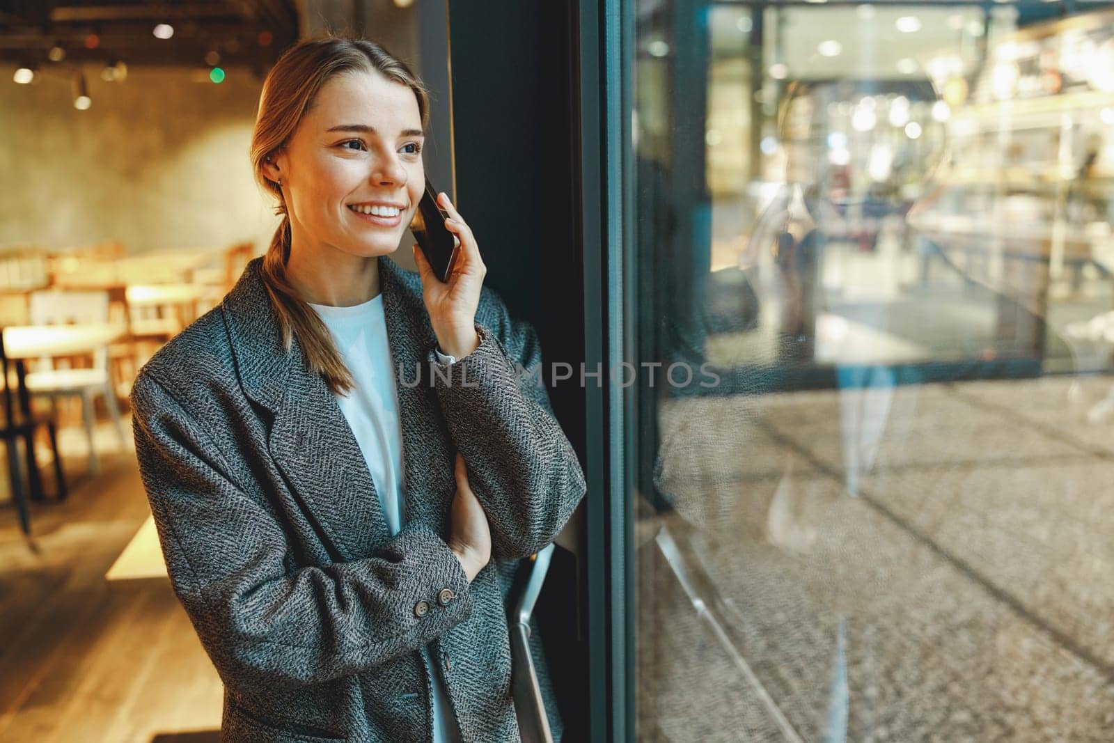 Stylish young female freelancer is talking phone with client while standing in coworking near window