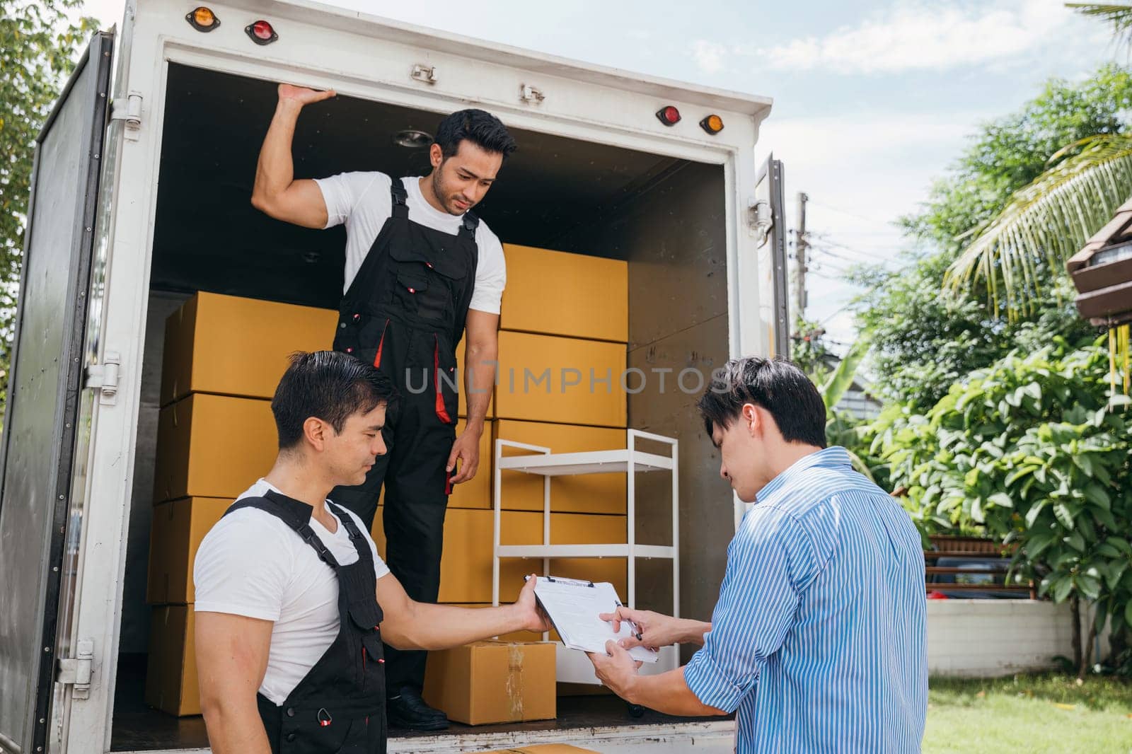 A couple pleased signs the delivery checklist after professional movers help with furniture lifting. Uniformed employees demonstrate teamwork for customer satisfaction. Moving Day Concept