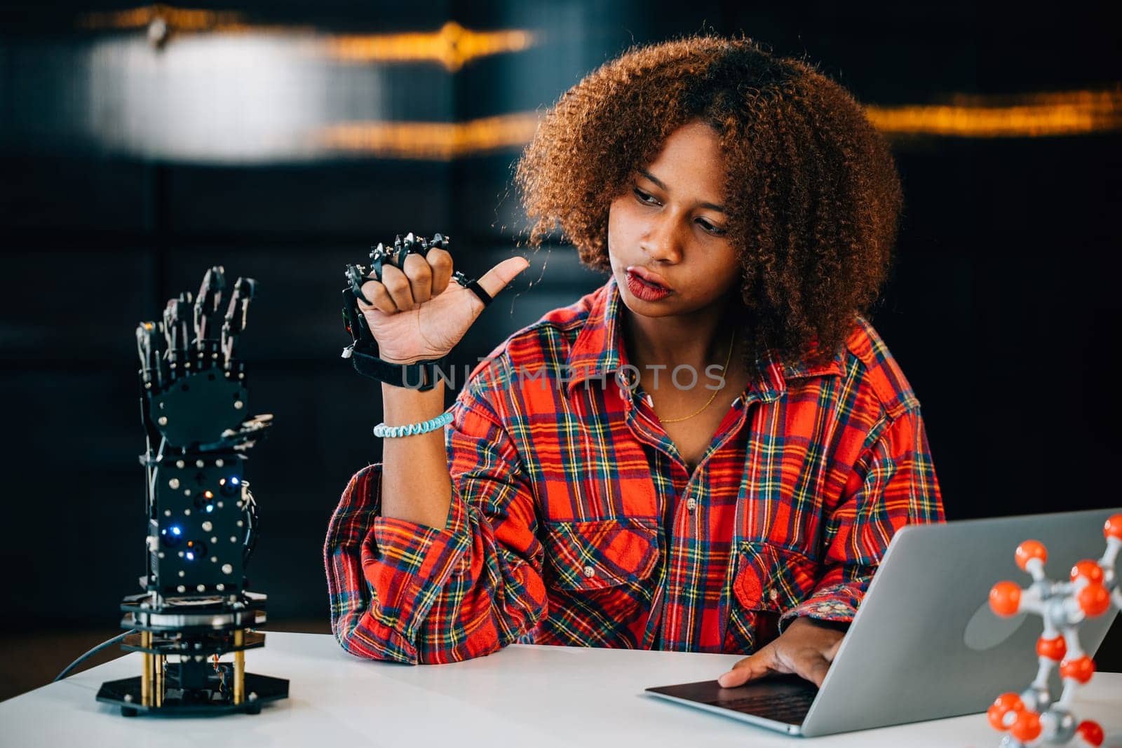 In a classroom a Black teenage student learns and controls a robotic arm for an engineering project. Displaying intelligence and skill she embraces innovation in technology. Robot hand. Innovation by Sorapop