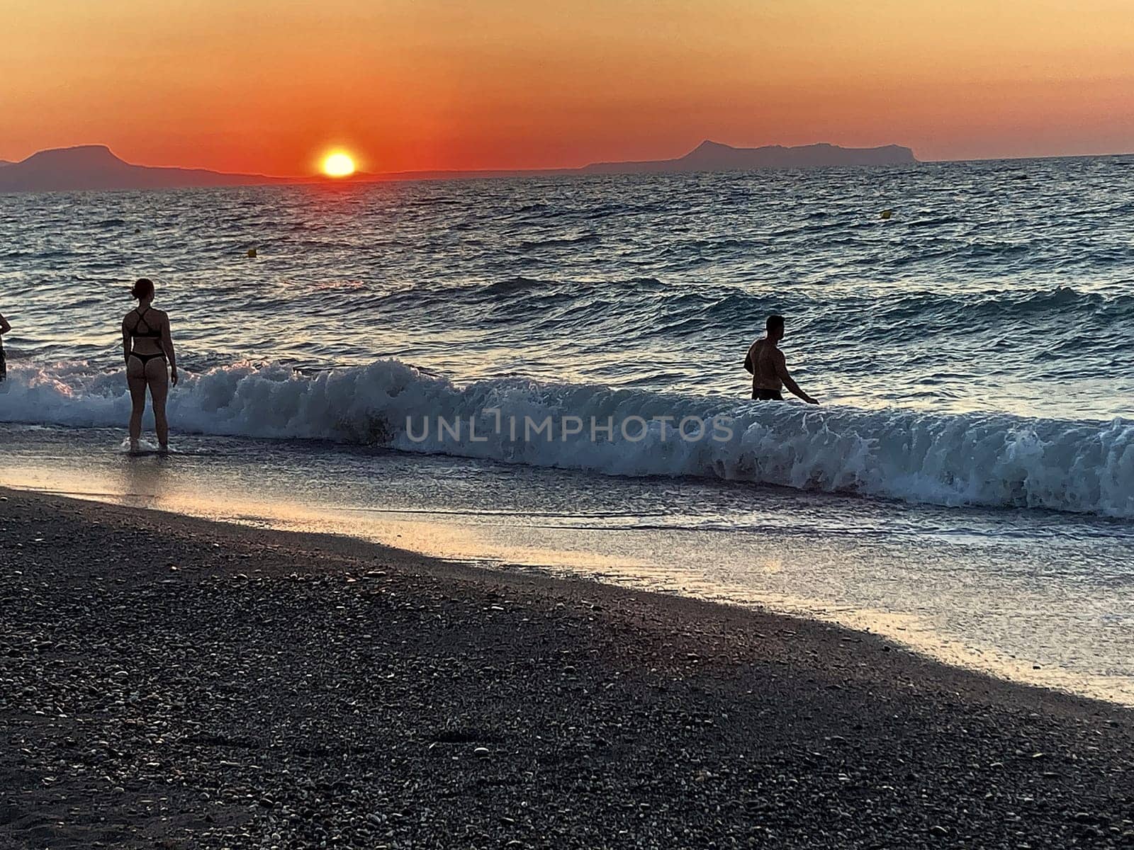 beautiful girl stands in the sea on the background of wonderful red sunset in Creta, Greece, 02 August 2023. young woman in black swimsuit staying by the waves of the sea water. Silhouette of a girl on the beach at sunset. by Costin