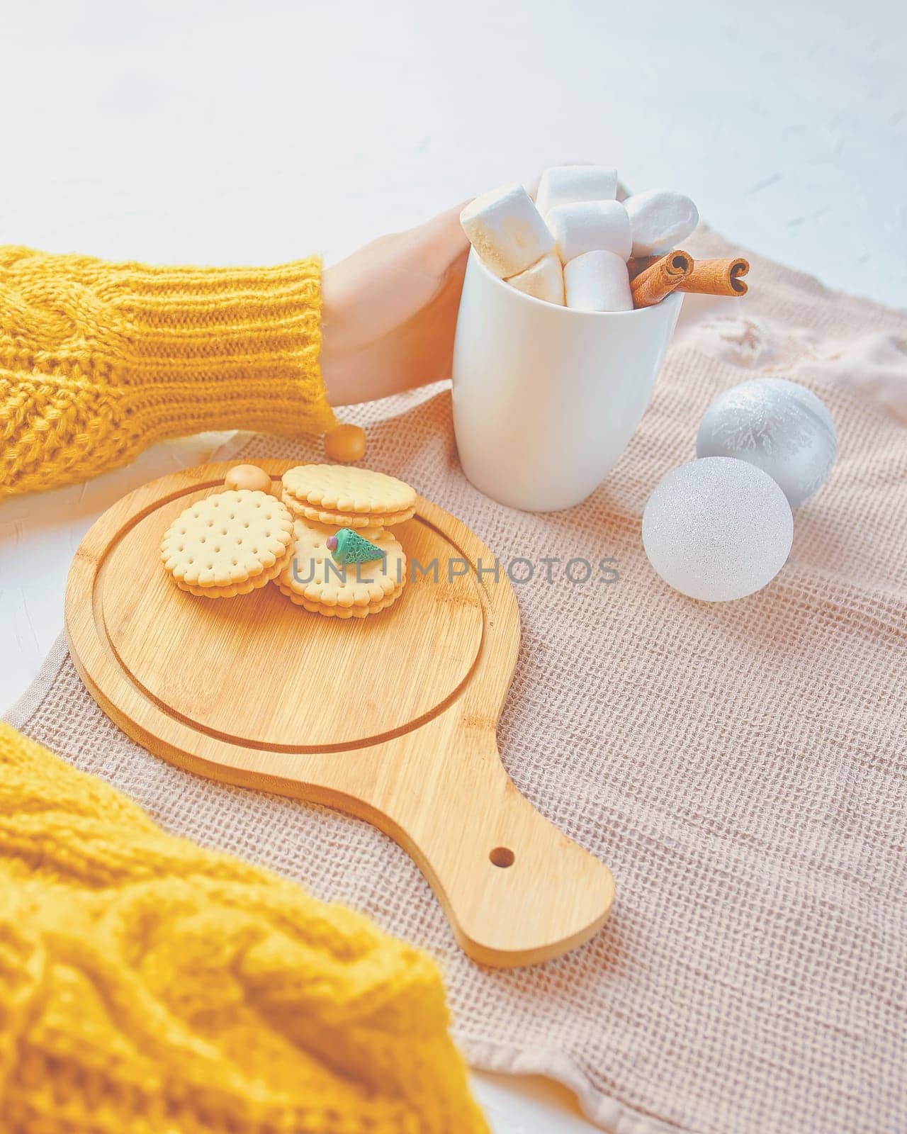 Woman in a yellow sweater drinks hot cocoa with marshmallow and cinnamon rolls in a white mug and cookies and a figure of a Christmas tree