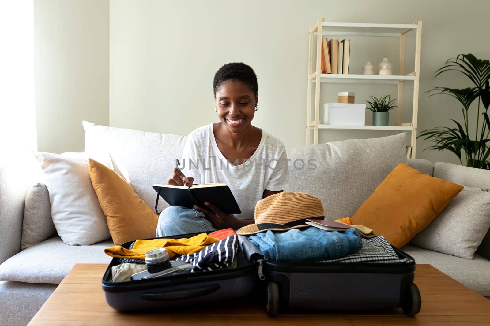 Young African American woman sitting on sofa checking packing list before summer holidays. Preparing suitcase for vacations. Vacation concept.