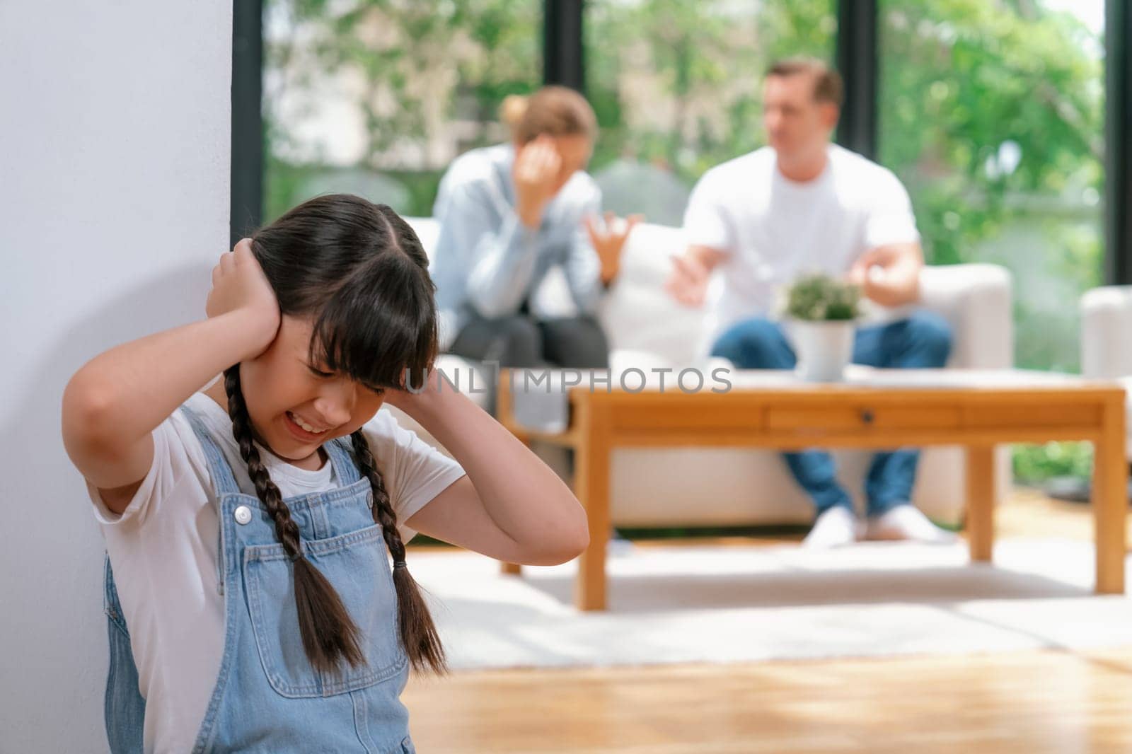 Stressed and unhappy girl huddle in corner, cover her ears with painful expression while her parent arguing in background. Domestic violence and traumatic childhood develop to depression. Synchronos