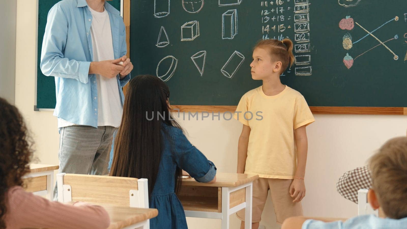 Professional teacher talking and explain idea to asian child during class. Caucasian student listening instructor while standing in front of classroom with blackboard math lesson written. Pedagogy.
