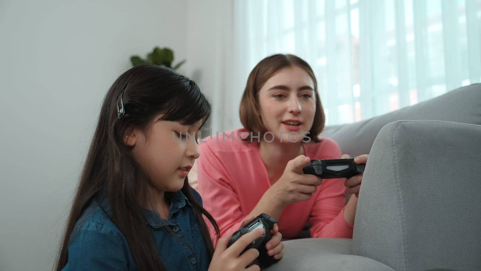 Happy girl playing game and sitting while mom lie on sofa by using laptop screen. Caucasian parent and attractive girl spend time together while holding joystick and focus on winning games. Pedagogy.