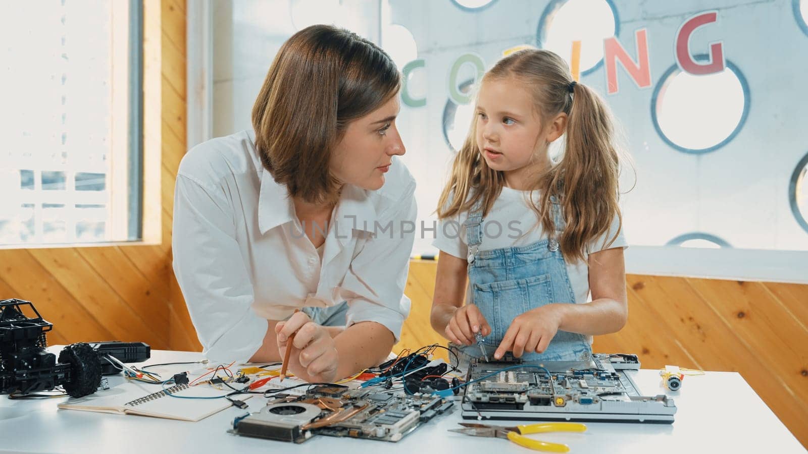 Young smart caucasian teacher teaching students about part of electronic board. Expert girl learn about digital electrical tool and fixing motherboard at table with chips and wires placed. Erudition.