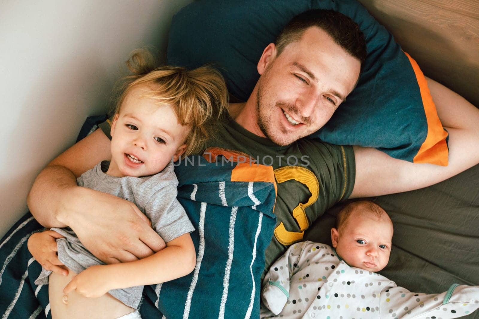 From above view of young handsome man in T-shirt sleeping in bed under blanket with little girl and baby