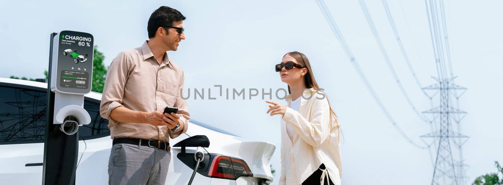 Couple pay for electricity with smartphone while recharge EV car battery at charging station connected to electrical power grid tower as industry for eco friendly car utilization. Panorama Expedient